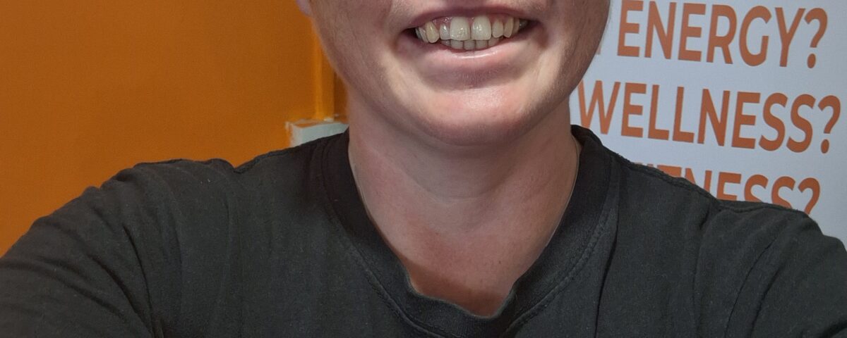 Corinna smiles in a selfie. She has orange hair and glasses, wearing a Fired Up Nutrition t-shirt.