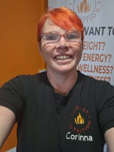 Corinna smiles in a selfie. She has orange hair and glasses, wearing a Fired Up Nutrition t-shirt.