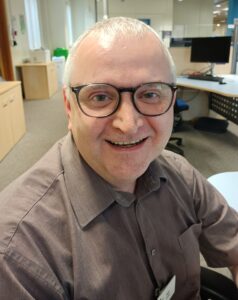 Nigel smiles at the camera, wearing round wide-rimmed glasses